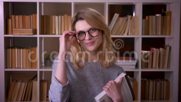 可爱的中年金发老师拿着一本书对着镜头微笑在图书馆修眼镜视频的预览图