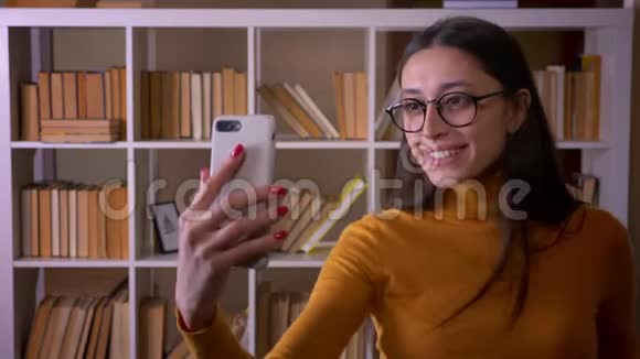 漂亮的黑发女教师戴着眼镜在图书馆的智能手机上做滑稽和奇怪的自拍视频的预览图