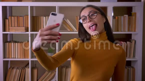 漂亮的深色头发的女老师戴着眼镜在智能手机上做滑稽的自拍在图书馆里欢快地露出舌头视频的预览图