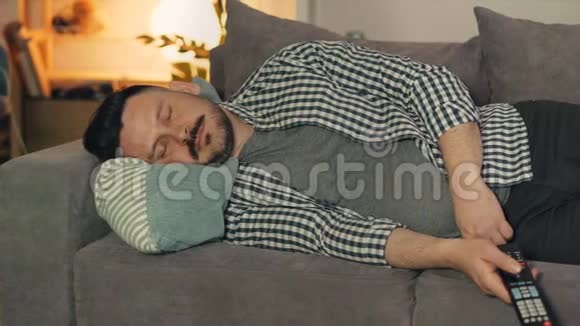 穿着休闲装的家伙在家里抱着电视遥控器睡沙发的画像视频的预览图