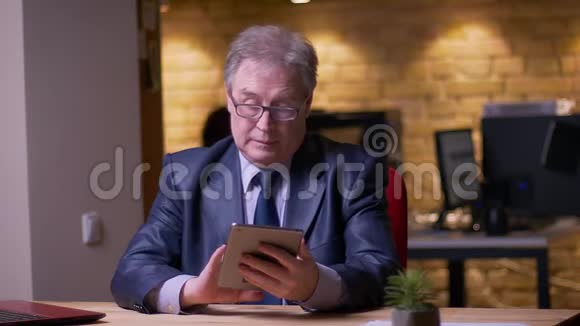 高级商人穿着正式服装坐在笔记本电脑前在办公室里处理平板电脑视频的预览图