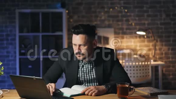 一个留胡子的人晚上在办公室用笔记本电脑在办公桌上用笔记本做笔记视频的预览图
