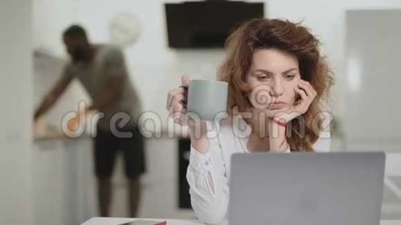 惊讶的白人妇女在开放式厨房的笔记本电脑上阅读新闻视频的预览图