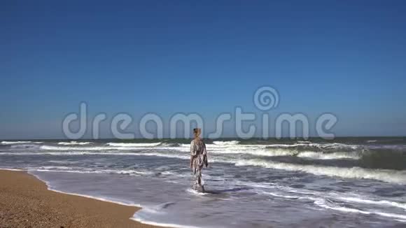一个身材苗条的女人穿着一件轻盈的长裙在夏日的海浪中沿着沙滩散步视频的预览图