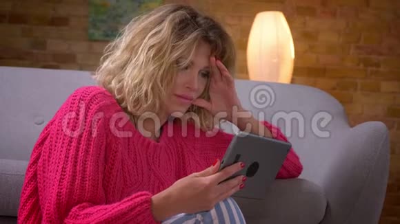 在舒适的家庭氛围中幸福的家庭主妇穿着粉红色毛衣观看平板电脑的特写镜头视频的预览图