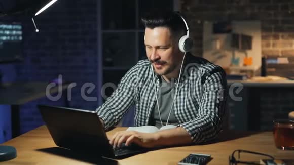 快乐的办公室工作人员戴着耳机听音乐晚上忙着用笔记本电脑视频的预览图