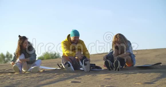 两个年轻的女人和一个男人朋友坐在沙丘上的滑雪板上穿着滑板设备靴子视频的预览图