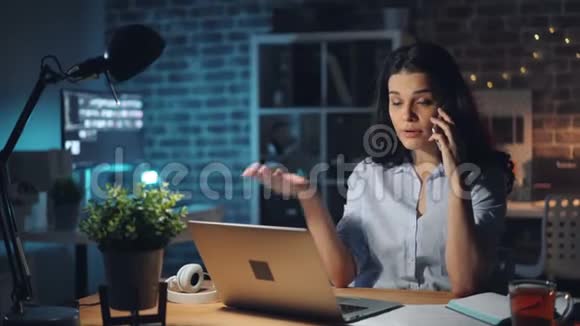 愤怒的年轻女士晚上在办公室用手机谈论问题视频的预览图
