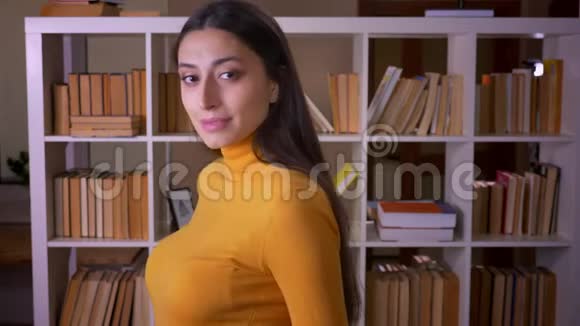 时尚的黑发女教师的肖像转向相机并在图书馆高兴地微笑视频的预览图