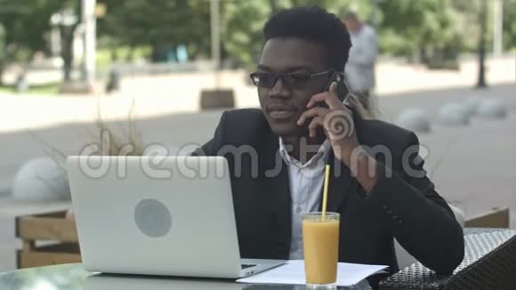 年轻迷人的美国黑人商人戴着眼镜和笔记本电脑坐在咖啡馆里用手机说话视频的预览图