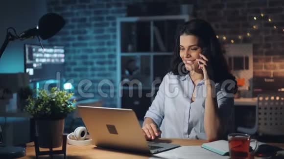 快乐的女人在黑暗的办公室里用手机和笔记本电脑聊天视频的预览图
