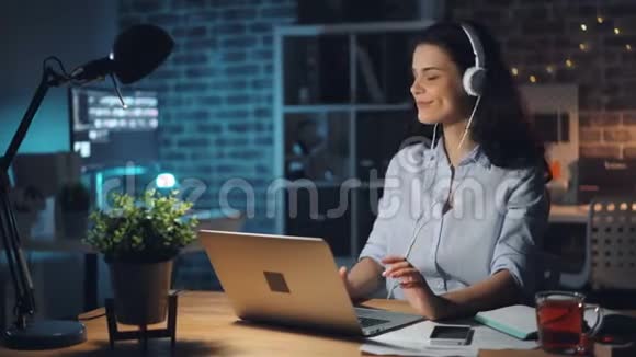 戴着耳机听音乐的漂亮女士在黑暗的办公室里用笔记本电脑工作视频的预览图