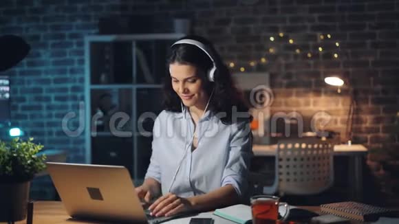 可爱的女孩用笔记本电脑工作晚上在办公室用耳机欣赏音乐视频的预览图