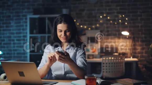 使用智能手机触摸屏幕的年轻女士在晚上加班视频的预览图