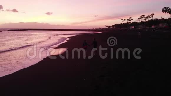 年轻的一家人在日落时沿着大海上的黑色海滩散步从无人机上观看特内里费西班牙视频的预览图