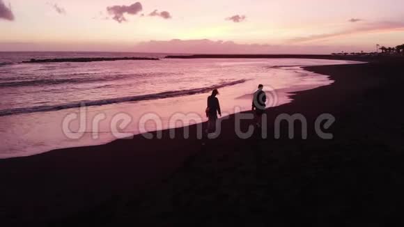 空中飞行一男一女在粉红色美丽的夕阳下沿着黑色的火山海滩奔跑特内里费西班牙视频的预览图