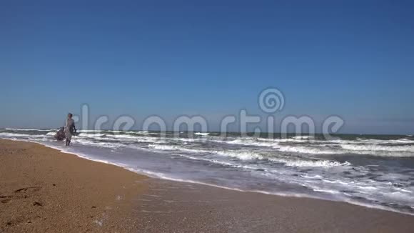 一个身材苗条的女人穿着一件轻盈的长裙在夏日的海浪中沿着沙滩散步视频的预览图