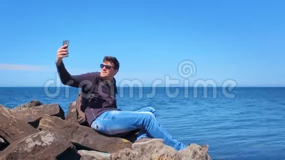 年轻人旅游博主在海上智能手机上自拍坐在石头上视频的预览图