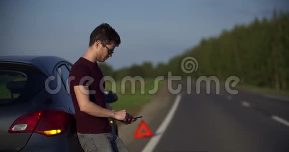 戴太阳镜的年轻人的肖像看着电话里的东西一辆破车在路上的车架上视频的预览图