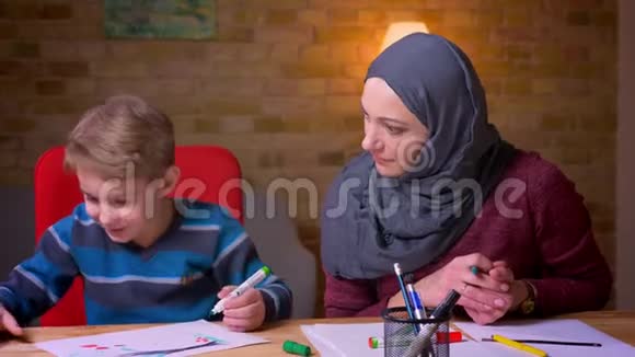 快乐的穆斯林母亲戴着头巾她的小儿子在舒适的家庭氛围中用记号笔画画视频的预览图