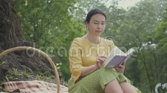 肖像可爱优雅美丽成熟的女人坐在公园树下的毯子上看书威克视频的预览图