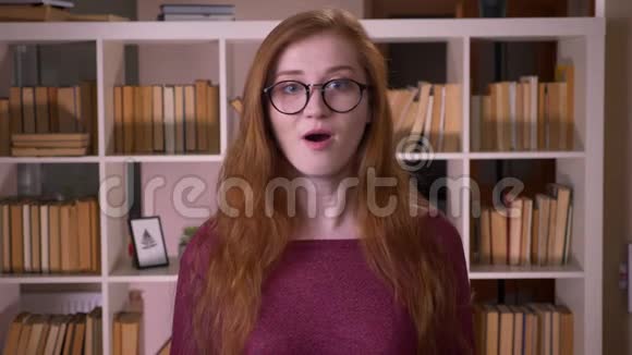 戴眼镜的年轻红发女子的特写肖像戴眼镜的白种人女学生兴奋而惊讶地看着视频的预览图