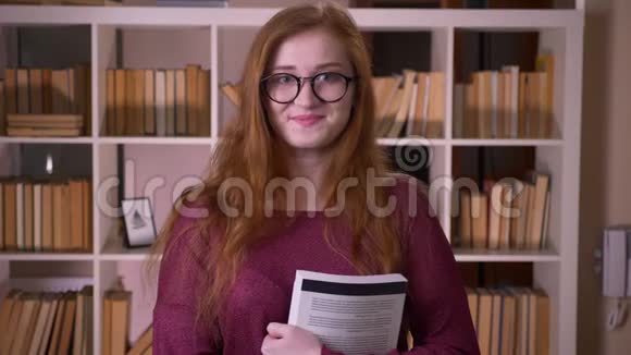 年轻的红头发迷人的白种人女学生戴着眼镜拿着一本书看着相机和视频的预览图