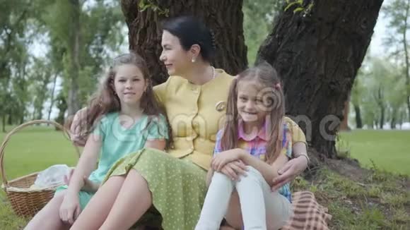 肖像可爱的成熟女人亲吻她的两个可爱的孙女坐在公园树下的草地上看着视频的预览图