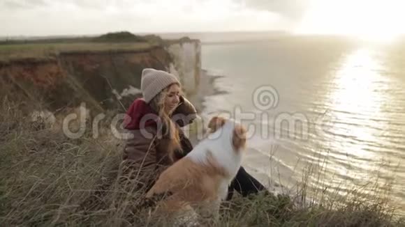 年轻的女孩子带着一只狗坐在诺曼底的岸边享受着令人愉快的日落吊车射击慢点视频的预览图