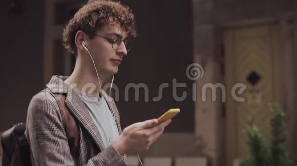 戴着检查夹克的戴着耳机戴着智能手机看屏幕的戴着眼镜的年轻微笑男人观看视频的预览图