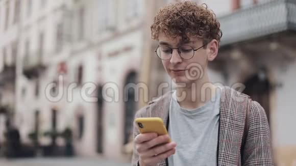 年轻的戴眼镜的学生使用他的现代手机设备与朋友聊天打字在屏幕上站立视频的预览图