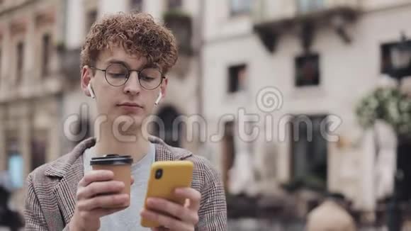 年轻的嬉皮士戴着眼镜和夹克带着耳机用他的智能手机交流和喝咖啡视频的预览图
