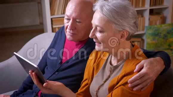 照片拍摄的是一对年长的白发白种人夫妇他们在舒适的家里和朋友一起在平板电脑上玩视频游戏视频的预览图