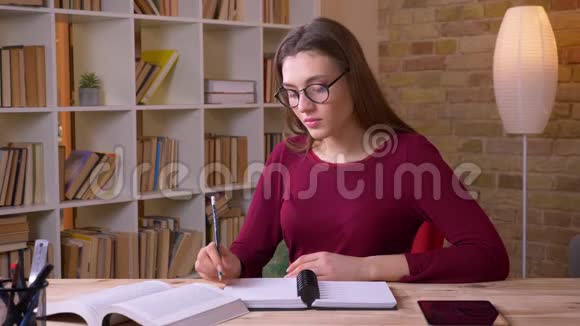 年轻的黑发女学生戴着眼镜在办公室里做笔记和手表进入相机视频的预览图