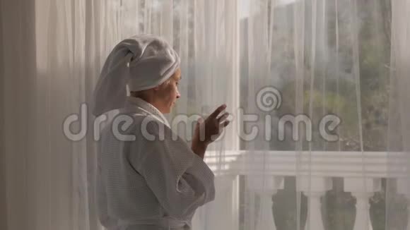 一个穿着白大褂头上拿着毛巾的女人从站在大窗户边的玻璃杯里喝水视频的预览图