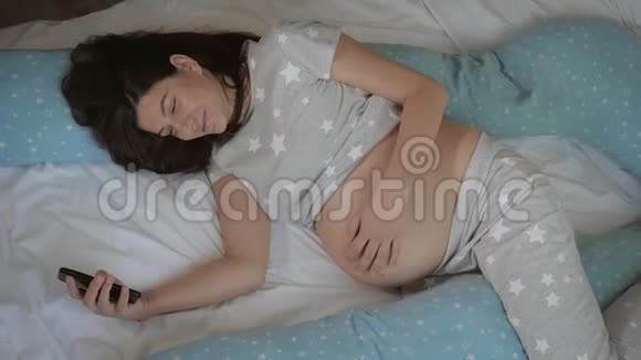 孕妇用智能手机躺在床上踩着她的胃视频的预览图