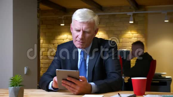 一个老白种人商人在平板电脑上浏览在办公室的室内喝咖啡的特写镜头视频的预览图