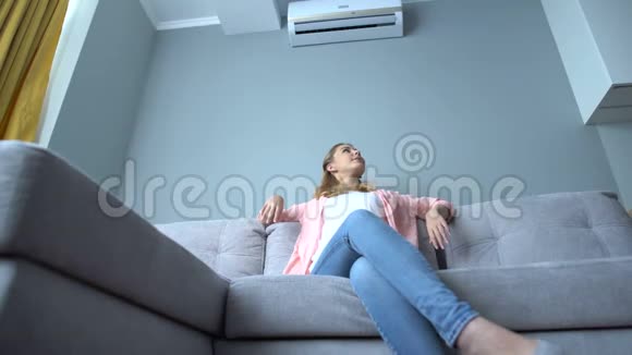 年轻女子坐在空调下的沙发上在炎热的夏天降温视频的预览图