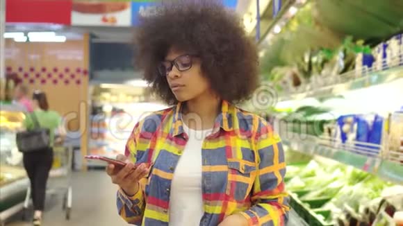 美丽的非洲裔美国妇女有着非洲发型手里拿着智能手机很难在超市里选择视频的预览图
