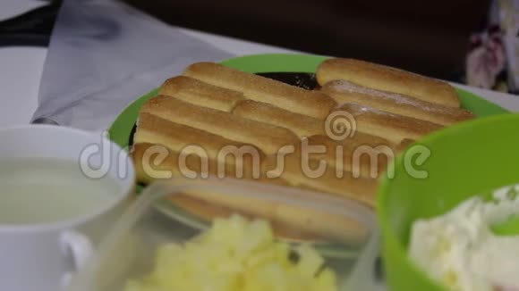 一个女人用萨沃亚迪饼干做蛋糕蘸上糖浆将饼干成排放置在基质上靠近菠萝和奶油视频的预览图