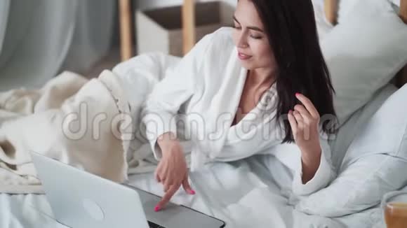 穿着浴袍的年轻女子躺在床上用笔记本电脑在网上工作或学习视频的预览图
