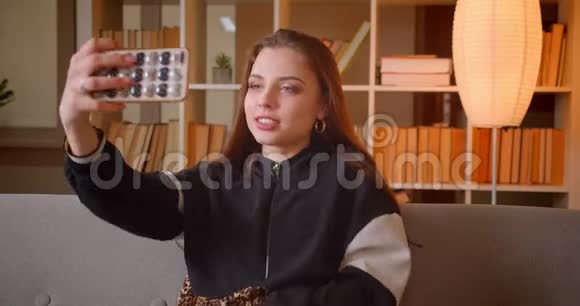 年轻少女的肖像制作手机自拍照片坐在沙发上书架背景在家视频的预览图
