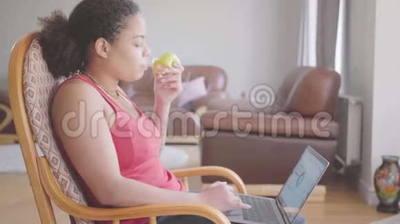 漂亮的非裔美国女人坐在扶手椅上一边吃苹果特写一边分析笔记本电脑上的图表快乐视频的预览图