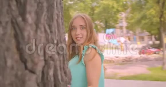 年轻可爱长发美丽的女性躲在树后看着镜头在镜头中露出漂亮的头发视频的预览图