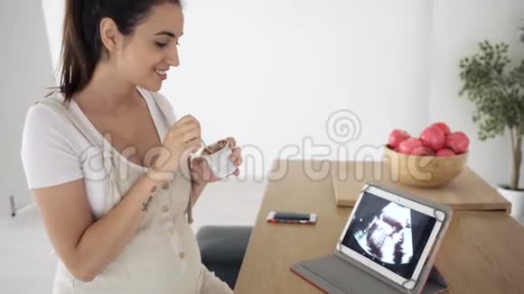 美丽的年轻孕妇在家里用数码平板电脑看婴儿的超声波时一边吃着冰淇淋视频的预览图