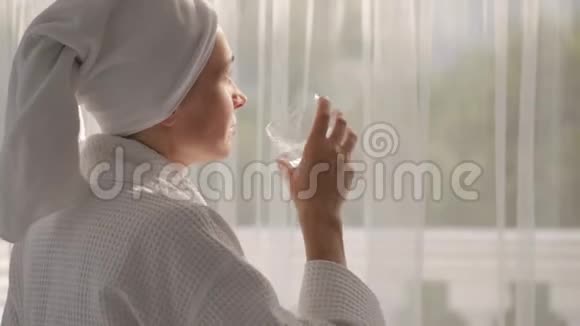 一个穿着白大褂头上拿着毛巾的女人从站在大窗户边的玻璃杯里喝水视频的预览图