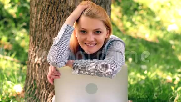 一个女孩坐在公园的草地上靠在笔记本电脑上看着相机一个学生在公园里休息视频的预览图