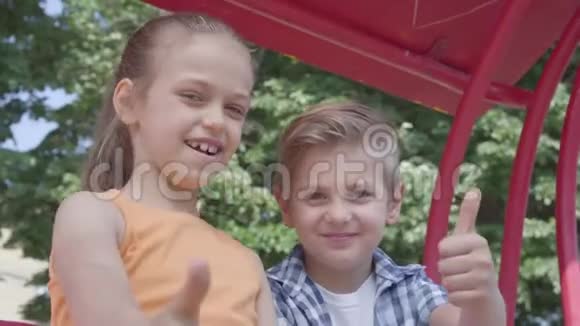 可爱的男孩和女孩竖起大拇指坐在公园里的秋千上看着镜头一对快乐视频的预览图