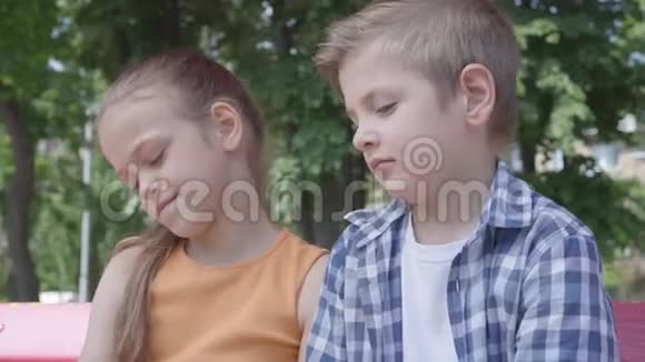 一个可爱的金发男孩在操场上坐在秋千上亲吻一个漂亮女孩的脸颊的肖像那个女孩很害羞A视频的预览图