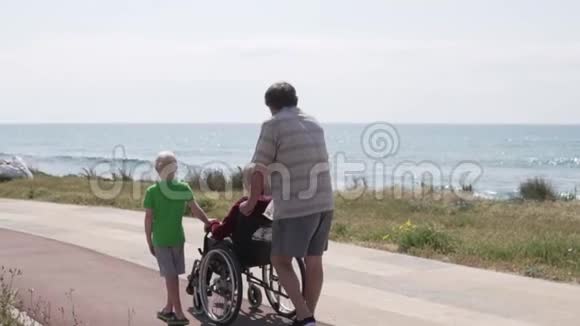 一个男人带着一个男孩带着轮椅和他的祖母沿着海岸走视频的预览图